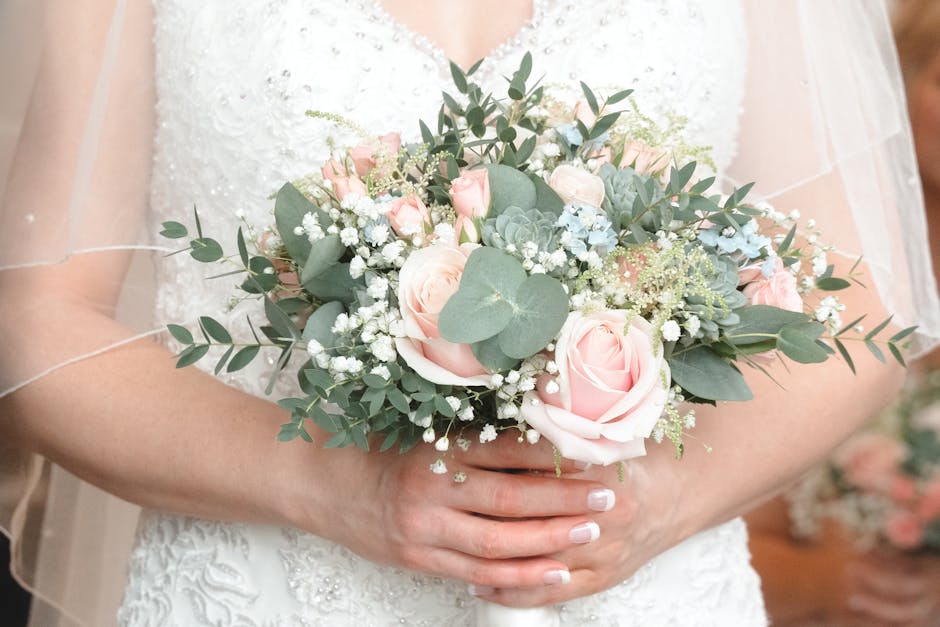 A person dressed in a white, textured wedding dress is holding a decorated floral bouquet. The bouquet features soft pink roses, sprigs of white baby's breath, green eucalyptus leaves, and succulent p