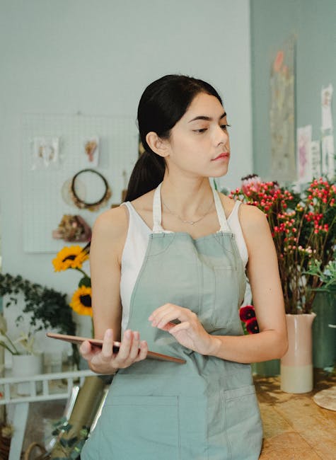 A young woman with long dark hair and light skin stands in a flower shop, wearing a light gray apron over a white sleeveless top. She holds a tablet in her left hand and appears to be considering flor