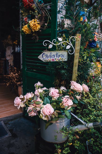 A display of fresh flowers in a floral shop or garden setting, featuring light pink roses with lush green leaves arranged in a metal watering can. Behind the flowers, there is a green wooden shutter w