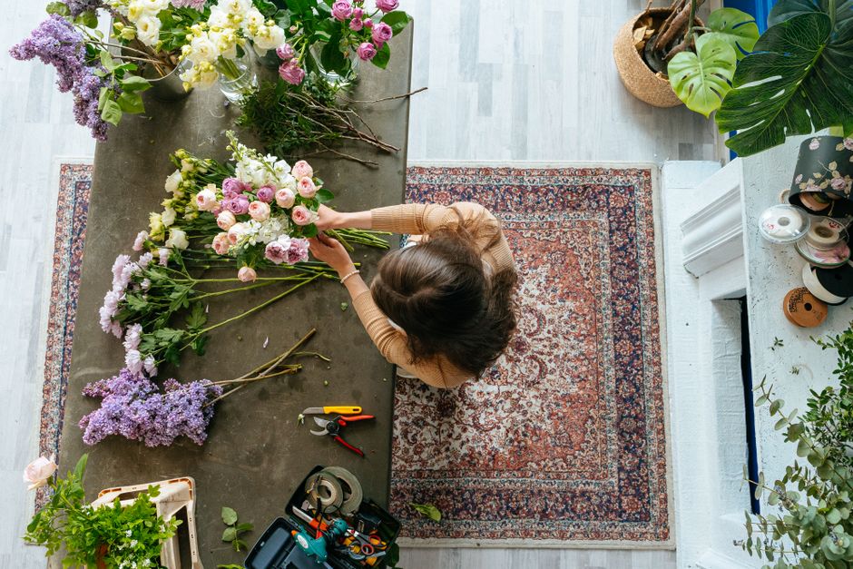 A woman arranging a bouquet of fresh flowers featuring pink ranunculus, white roses, and purple larkspur on a dark wooden table in a well-lit, cozy indoor space. The table is scattered with flower ste