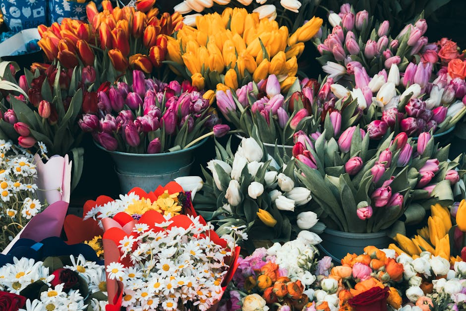 A vibrant display of fresh flowers including pink, white, purple, and orange tulips arranged in metal buckets and wrapped bouquets. The tulips have smooth, slightly curved petals with some opening to 